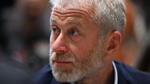 A close of up a bearded Roman Abramovich in a blue suit and white shirt looking to his right