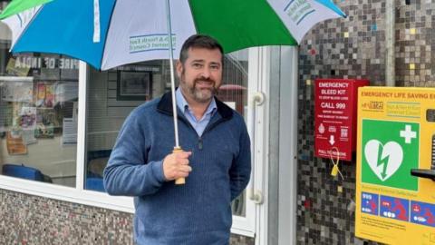 Councillor Kevin Guy with a newly-installed bleed kit at Community at 67 in Keynsham. He is standing next to the kit, which has been installed on a stone wall. He is wearing a blue jumper and holding an umbrella.