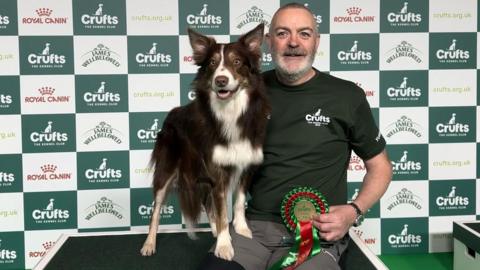 Mark Douglas and his dog with the winning rosette, which is coloured red and green. They are sitting on a podium, in front of a Crufts advertising hoarding. Mark is wearing a dark green polo shirt and grey trousers. He is smiling and holding the dog on his lap. The dog is looking into the camera, it is alert and has its mouth slightly open.