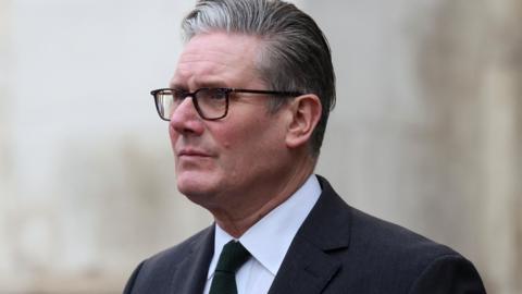 Sir Keir Starmer looking off to the left hand side of the frame, wearing dark brown tinged glasses. He is wearing a dark suit and a green tie.
