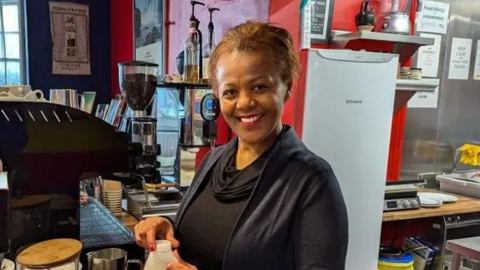 Tutu Melaku is standing in her kitchen smiling and opening a carton of milk. A fridge and a coffee machine is situated behind her.