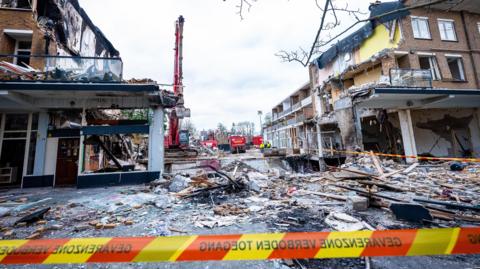 A damage apartment block with a wide gap in the middle where the explosion took place. There is rubble an wood on the ground.  In the background is a large digger. In the foreground is a strip of emergency crew tape.