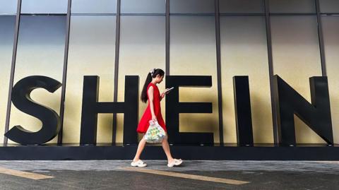 A woman in a red dress looks at her smartphone while walking past a Shein sign outside the company's office in Guangzhou, China. 