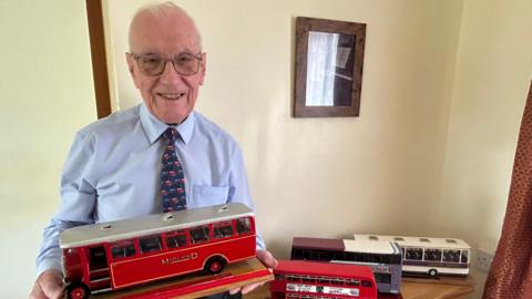 Arthur Mansell has white hair and glasses. He is wearing a blue shirt and blue and red tie. He is holding a red replica bus model. On a table next to him are three replica buses.