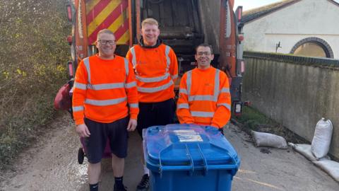 Three men standing behind a bin lorry. They are smiling and wearing bright orange jumpers that are hi vis. There is a blue wheelie bin in front of them.