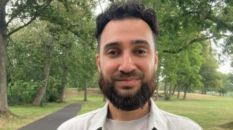 Fahad Khan wearing a beige tshirt and shirt standing in a park 