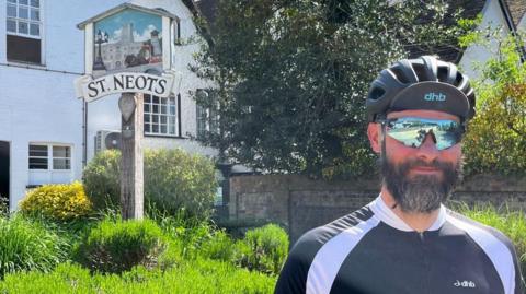 Richard Slade is standing in front of a grassy backdrop with the town sign of St Neots behind him. He is wearing a tight-fitting blue and white cycling top, a bicycle helmet and has sunglasses on. He has a greying beard and moustache.