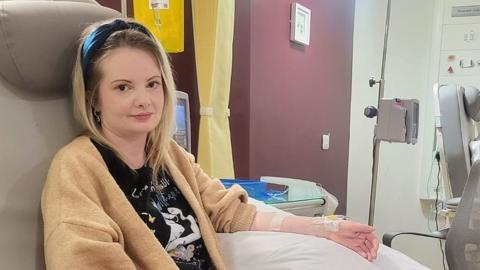 Gemma Coleman with long blond hair wearing a black headband and black t-shirt with a gold coloured cardigan sits in a medical room. Her sleeve on her left arm is rolled up and she has a drip going into her wrist. She is smiling.