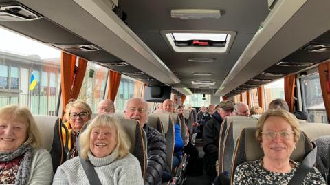 A group of people siting inside a bus with their seat belts fastened. They are all smiling at the camera. 