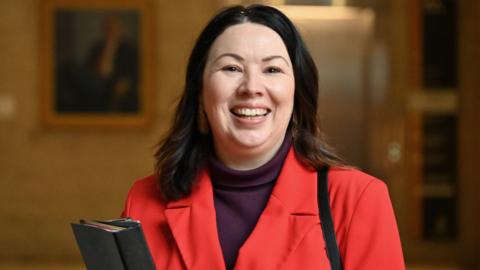A smiling Monica Lennon holding a dark ring binder. She is wearing a purple turtle neck and a bright red jacket.