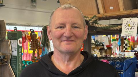 Man looking into camera, wearing black jumper and standing in pet food shop.