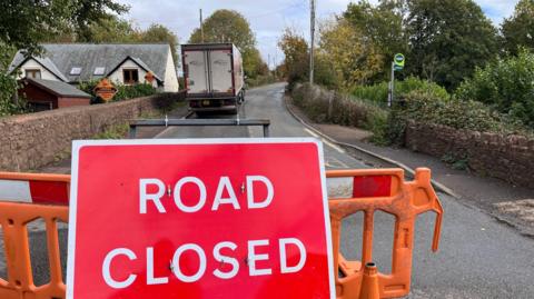 Big red road closed sign. There's roadworks barrier stopping people from driving across the bridge in front.