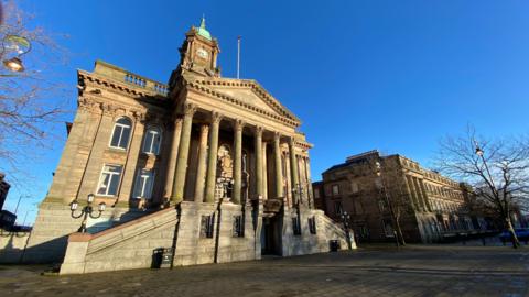 An historic building with classical architecture, featuring a wide stone staircase leading to its entrance. It has an ornate clock tower with a flagpole. In front of the building is a paved courtyard with trees.