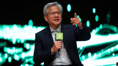 Jensen Huang is speaking into a microphone and sitting in front of a bright green and white lit background 
