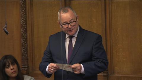 A bespectacled man in a navy suit, dark red tie and white shirt stands and reads off a piece of paper.