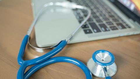 A picture of a blue and silver stethoscope placed upon an Apple Mac laptop computer, placed on a wooden desk.