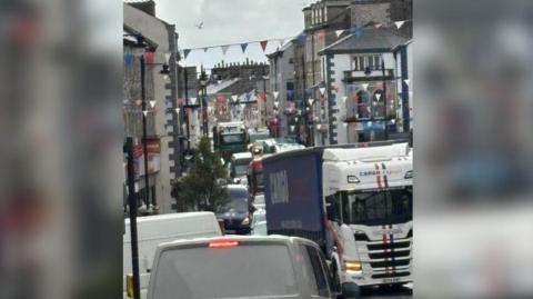 Kendal Highgate (the town's High Street). It is gridlocked with traffic, with large HGVs in the small road. There are cars parked the opposite direction on a one way street. There are market flags and shops on either side of the road, but this is obscured with how busy the road is.