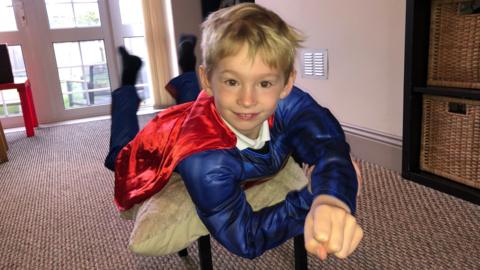 Fletcher is wearing the Superman costume whilst suspended on a chair, pretending to fly.