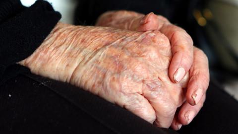 An elderly person's hands that rest in their lap. They wear pink nail varnish which has slightly chipped.