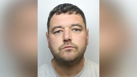 A man in his thirties, pictured in a police mugshot. He has short, mousy brown hair, blue eyes and stubble. He is wearing a grey t-shirt.