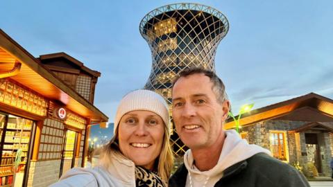 A blonde woman wearing a white woolly beanie and a white jacket and patterned scarf. She is standing next to a man, with greying hair, who is wearing a black coat and white hoodie. They are standing in front of a monument.
