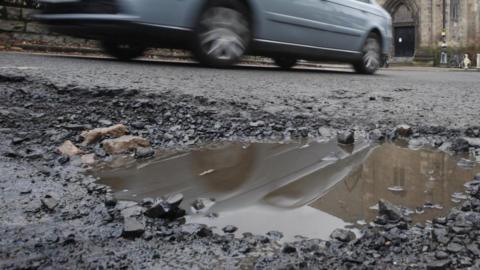 A ground-level image of a pothole in a road, full of water. A light blue car is driving past in the background. The entrance to a building is also visible in the distance.
