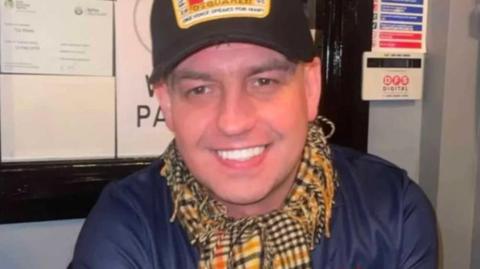 A man wearing a cap with a canadian flag on it, he is wearing a yellow check scarf. He is smiling