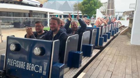 Adult men and women sit two by two in navy blue carriages of a rollercoaster which is stopped at the platform. They are smiling and clapping. 