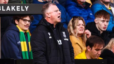 Stoke City manager Mark Robins, standing in front of Norwich City fans, shouts instructions to his players