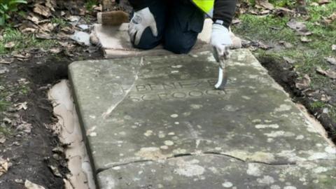 A person is kneeling above a gravestone that is lying in the ground. The stone has cracks over it. The person is using a thin brush to clean inside letters that read "Ebenezer Scrooge"