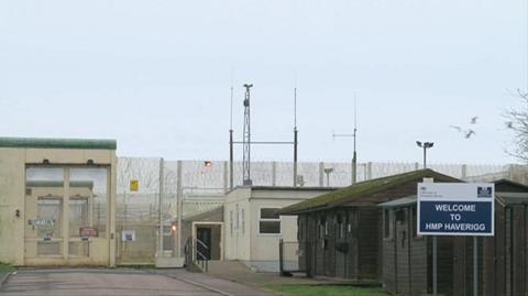 The entrance to HMP Haverigg in Cumbria. There is a blue and white sign reading Welcome to HMP Haverigg. The area is fences off by a tall cream fence with wire along the top and warning signs on the gate. Two small hut-type buildings painted brown are outside the gates on one side of the entrance.