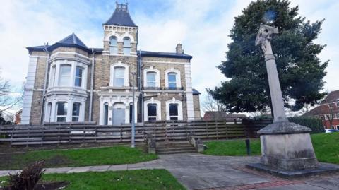 The two-storey villa with a turret has white, arched windows. In front there is a wooden fence with steps leading down a stone monument. 