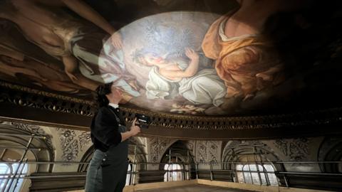 A woman shines a torch on the painting on the Blenheim Palace Great Hall ceiling.