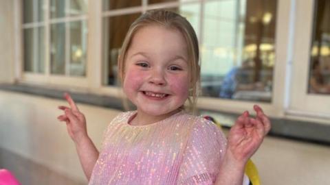 Brie McCann, a 5 year old girl with blond hair, smiling for the camera while wearing a pink dress