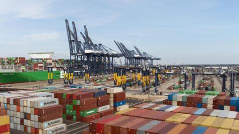 Shipping containers are at the front of the image, in the background are blue cargo cranes with a green ship moored on the left.