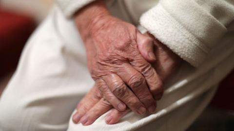 An elderly person rests their hands over one another on their lap. They wear white trousers with a white long sleeved jumper.