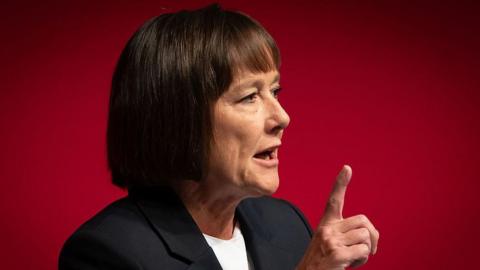 Llun yn dangos dynes efor gwallt cwta yn pwyntio bys i fyny, o flaen cefndir coch. Mae hi'n gwisgo siwt ac efo'i cheg ar agor fel ei bod hi yng nghanol siarad.