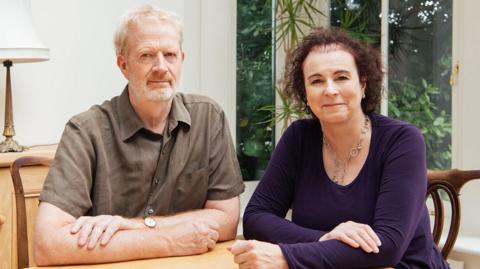 Charles (left) and Liz Ritchie are seated at a wooden table in a bright room with large windows. They both have their arms resting on the table. Charles has receding grey hair and a thin beard, he is wearing a brown short-sleeved shirt. Liz has thinning brown curly hair and is wearing a purple top and silver hooped chain around her neck. Behind them is a lamp on a side table, and a large window from which tall green plants are visible.
