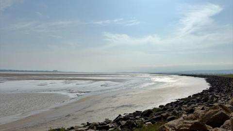 A sweeping coastline stretches out in a semi circle, with the sea so far out it is hardly visible. Large rocks go as far as the eye can see in the right of the photo, with the rocks then meeting the golden sand. The sand appears to be damp with the sunlight shimmering off of it and there is a small pool of water in the right of the image. The sky is blue with a few small clouds.