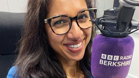 A smiling Rena Annobil in the studio in front of a large purple microphone bearing the 91ȱ Radio Berkshire logo