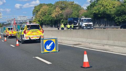 The scene of a fatal crash on the M20. There are two National Highways vehicles and a series of cones closing off the stretch of motorway. There is a tow truck on the other side of the motorway.