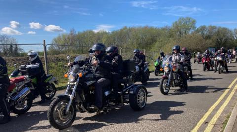 A large groups of trikes and bikes make their way down a narrow road on a sunny day.