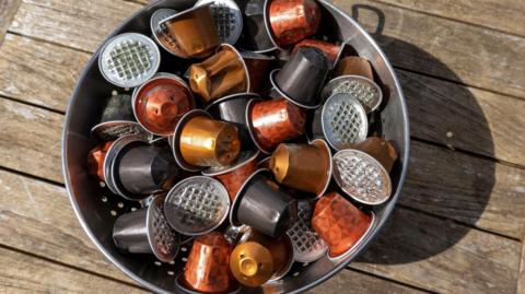 Used coffee capsules ready for the aluminium tops to be recycled. They are collected in a bin that is placed on top of a wooden surface outside on a sunny day