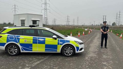 A police car is parked on a roadside, a uniformed police officer is standing next to it, the road has traffic cones along either side and in the distance are electricity pylons