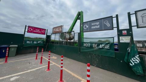 The Hempsted Household Recycling Centre. Bays for 'small electricals', 'wood and timber', 'scrap metals', and 'general waste'. Orange and white striped bollards sit in front of them.