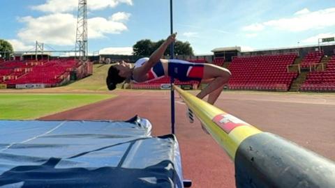 Emily MacDonald high-jumping over a bar, her body parallel to the ground as she clears, in the middle of an athletics stadium surrounded by rows of empty, red seats. A blue landing mat underneath the bar awaits her descent.