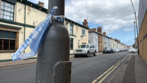 A close-up of part of a torn police cordon tied around a black lamppost. I the background are houses and cars parked at the side of a road.