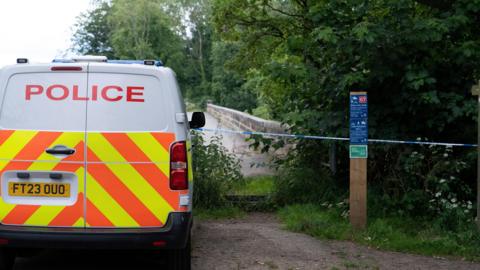 A police van is parked facing away from the viewer, it is parked behind a blue and white police tape. A bridlepath sign on a wooden post is to the right of the van.