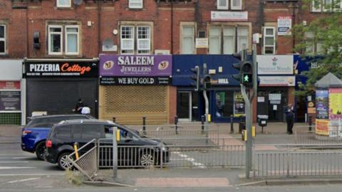 A daylight scene of a row of shops on a street. One shop has a black and orange sign which reads 'Pizzeria Cottage'. Next door is another shop with a sign which has silver writing on a purple backdrop and reads 'Saleem Jewellers'. In the foreground are cars on the road and a pedestrian crossing. 