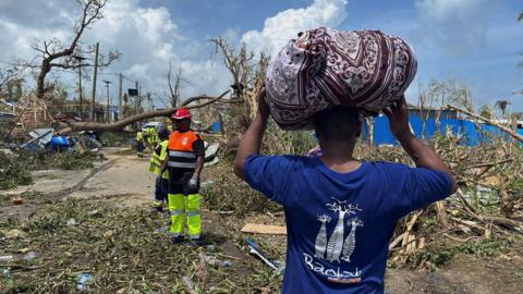 A man carries his belongings on his head while rescue workers sift through the debris.